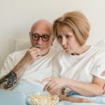 Senior couple bonding over movie night with popcorn and laptop in cozy home setting.